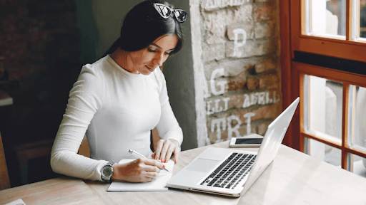 Focused woman writing notes while using a laptop in a cafe